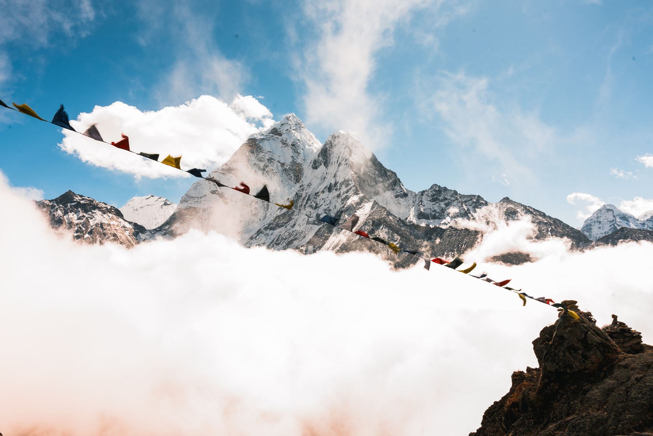 Ein verschneiter Himalaya-Gipfel ragt über eine Wolkendecke, davor sind bunte tibetische Gebetsfahnen gespannt.